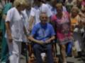 Una persona en silla de ruedas es acompañada por varios adultos, algunos de ellos vestidos con uniformes blancos, en un entorno urbano. La escena parece ser una visita o una reunión de salud, con personas de diferentes edades y estilos de vestimenta.