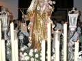 Estatua religiosa de una Virgen con el Niño, rodeada de velas y flores en un ambiente festivo.