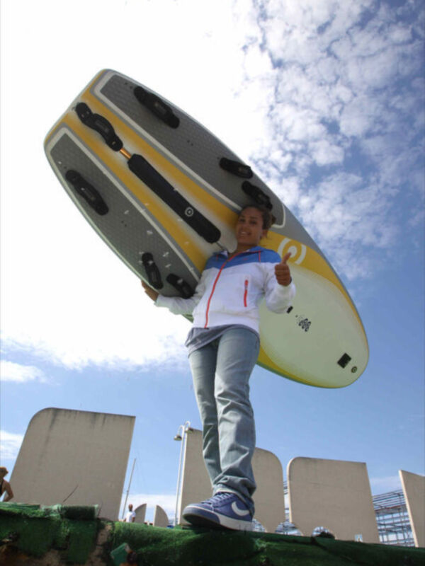 Una persona sostiene una tabla de surf gigante, con un cielo azul y nubes blancas en el fondo.