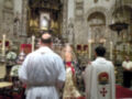 Imagen de dos sacerdotes en una iglesia, uno con un hábito blanco y otro con un hábito rojo y dorado. En el fondo, se ve una imagen de la Virgen María en un altar con velas y decoraciones.