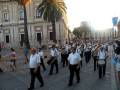 Desfile militar en una plaza con palmeras y edificio histórico. Músicos tocan instrumentos militares en uniforme blanco y negro.