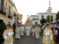 Ceremonia religiosa con personas vestidas de blanco y dorado, llevando estandartes y procesionando por una calle.