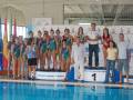 Equipo femenino de natación posando en una piscina con banderas y logotipos.