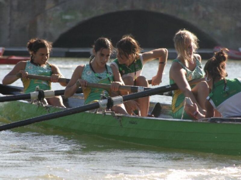 Equipo de remo femenino en acción en un barco verde, con un puente de fondo.