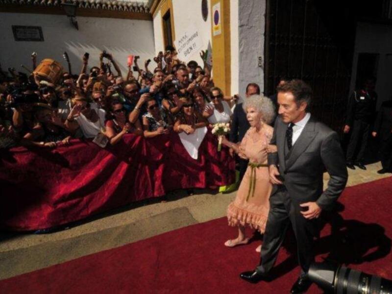Una celebridad femenina y masculina caminan por un camino de alfombra roja, rodeados por fotógrafos y asistentes. La celebridad femenina lleva un vestido largo de color rojo con detalles dorados, mientras que la masculina está en traje negro. La escena se desarrolla frente a una pared blanca con detalles arquitectónicos.