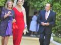 Imagen de una mujer vestida con un vestido rojo y tocado, caminando junto a otros dos individuos. El entorno parece ser un jardín o parque con vegetación en el fondo.