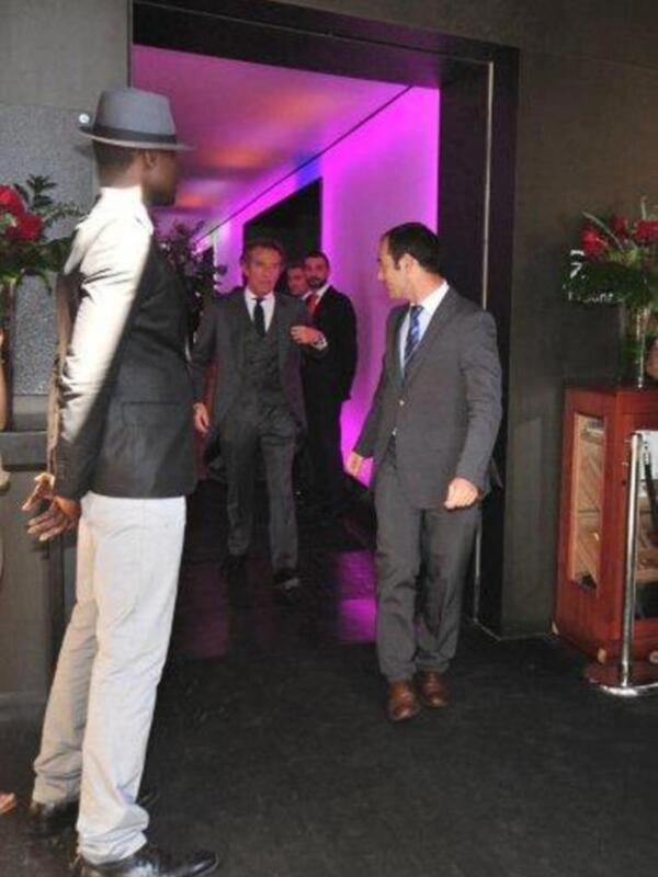 Un hombre en traje se dirige a un grupo de hombres en traje, todos con corbatas. La escena parece ser una reunión formal o evento empresarial en un ambiente elegante con luces de neón rosa y roja.