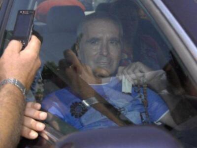 Un hombre en una camiseta azul está sentado en el asiento del conductor de un vehículo, mientras que otra persona le toma una foto con su teléfono móvil.
