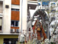 Una estatua religiosa en un arco de metal, rodeada por candelabros y flores. El fondo es una calle con edificios modernos, uno de los cuales tiene un balcón abierto donde se puede ver a una persona. La escena parece ser de una procesión o celebración religiosa en un entorno urbano.