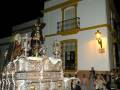 Fotografía nocturna de una procesión religiosa con una imagen de Cristo en la cruz, rodeada de luces y acompañada por fieles en la calle. La imagen se encuentra en una calle con edificios de estilo colonial, iluminada por faroles y luces de la calle.