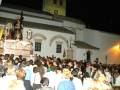 Una procesión nocturna con una imagen religiosa en el centro, rodeada de fieles que observan atentamente. La iglesia es visible con detalles arquitectónicos y una cruz en el frontis, iluminada por la luz de las farolas.