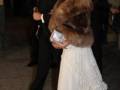 Una mujer en un vestido blanco con volantes y una chaqueta de piel marrón, posa junto a un hombre en traje negro. Ambos están en una alfombra roja, posiblemente en un evento formal.