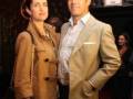 Una pareja posa juntos en un evento formal, ambos con trajes de estilo vintage. La mujer lleva una chaqueta beige y el hombre un blazer claro con camisa blanca. La escena sugiere una celebración o premiación.