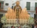 Procesión religiosa con estandarte de la Virgen, adornado con velas y flores, en una plaza con edificios blancos.