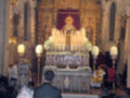 Imagen de una iglesia con un altar central adornado con velas y flores. Algunos fieles están presentes, incluyendo un sacerdote en el centro. La decoración es elaborada con detalles dorados y rojos, y hay una imagen central en el altar. La atmósfera es reverente y solemnemente festiva, típica de una celebración religiosa.