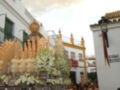 Procesión religiosa en una calle de la ciudad, con una imagen procesional en el centro, rodeada de flores y velas, y una bandera española en la parte superior derecha.