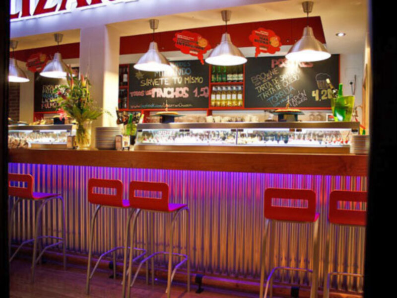 Interior de un restaurante con barra, sillas rojas y menú en la pared.