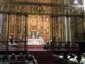Interior de una catedral con altar y retablo dorado, rodeado por fieles en bancos de madera.