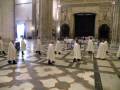 Los sacerdotes vestidos de blanco se alinean en una iglesia con arcos y columnas, preparándose para oficiar un servicio religioso.