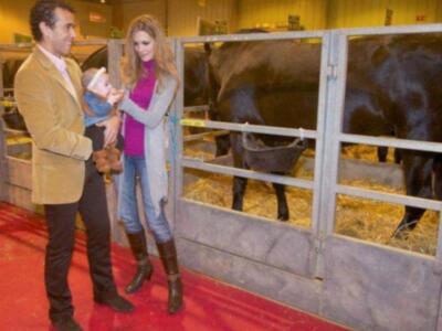 Una persona adulta y una niña están en un ambiente de exposición, posiblemente una feria o evento agrícola. La niña sostiene un bebé mientras la persona adulta la abraza. En el fondo, hay una granja con un animal de gran tamaño, posiblemente un toro o una vaca, dentro de una jaula con paja. El ambiente es iluminado y parece ser una exposición o evento público.