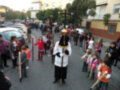 Una procesión religiosa en un barrio con personas vestidas de diferentes colores, algunos portando cruces y otros niños con vestimentas tradicionales. La imagen muestra una atmósfera festiva y cultural, con edificios residenciales al fondo.