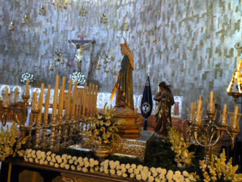 En una iglesia, se encuentra un altar adornado con velas encendidas y flores blancas. En el centro, una estatua de la Virgen María se eleva sobre un pedestal dorado. A su lado, una figura humana con sombrero y vestimenta colonial se apoya en un pedestal. La pared detrás del altar está decorada con una gran imagen de la Virgen María, iluminada por pequeñas luces doradas.