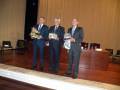 Tres hombres en trajes sostienen trofeos, posiblemente durante una ceremonia de premiación.