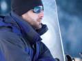 Un hombre con una chaqueta azul y un gorro negro, posando con una tabla de snowboard. El fondo es nevado y el cielo está oscuro, sugiriendo una tarde o noche de invierno.
