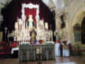 Imagen de una iglesia con un altar adornado con flores y velas. Un sacerdote y una persona vestida de blanco se encuentran frente al altar, rodeados por decoraciones religiosas. La imagen transmite una atmósfera de reverencia y devoción.