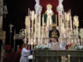 Imagen de una procesión religiosa con una Virgen en el centro, rodeada por velas y flores. La gente está vestida formalmente, con un altar al fondo.