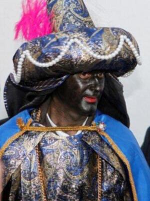 Un hombre con una expresión seria, vistiendo un traje tradicional con un sombrero azul ornamental y una corona de plumas rojas. La expresión sugiere seriedad y concentración.