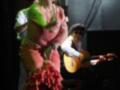 Una mujer vestida con un traje de flamenco colorido y verde, posando en una escena teatral. Detrás de ella, un hombre toca la guitarra mientras otra persona observa el espectáculo.