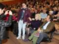 Una persona joven toma una foto de una anciana sentada en el público durante un evento. La anciana lleva una chaqueta de terciopelo y un par de medias de encaje. La audiencia parece estar disfrutando de la presentación.