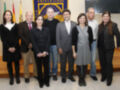 Grupo de personas en una reunión formal, posando para la foto. La imagen incluye a varios individuos vestidos con ropa formal, posando frente a una pared con banderas y un cartel de la Universidad. La escena parece ser en un entorno institucional o académico.