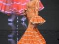 Una modelo presenta un vestido de flamenco naranja con volantes en las mangas y falda, destacando la cultura española.