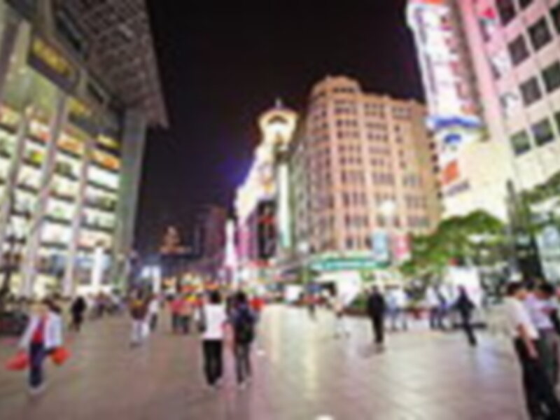 Área urbana nocturna con edificios modernos y luces brillantes, con personas caminando en el centro.
