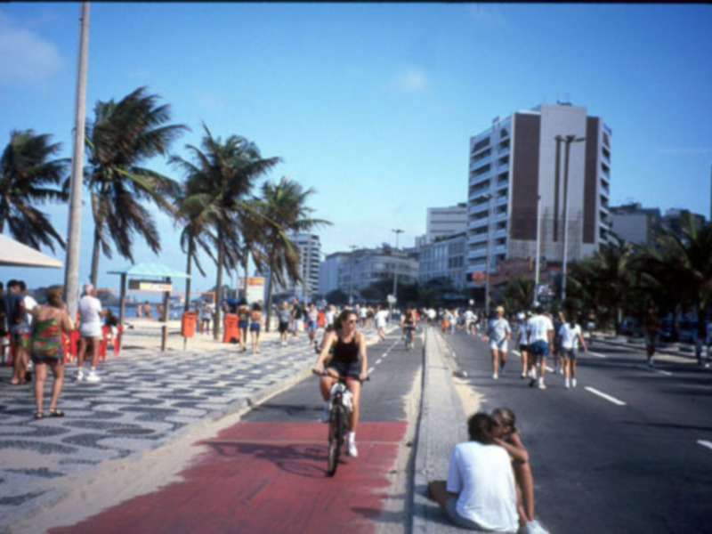 Una mujer en bicicleta se mueve por la acera, mientras otros pasean o esperan en el borde de la calle. El horizonte muestra edificios altos y árboles, con un cielo claro.