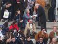 Un hombre en traje negro y corbata se inclina para saludar a otro hombre con una camisa roja. Ambos están en un entorno público, posiblemente un estadio o plaza, con personas sentadas y otros en pie alrededor. La escena sugiere una interacción formal o de bienvenida entre los dos individuos.