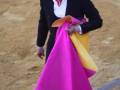 Un torero en el ruedo, con un traje negro y una camisa blanca. Lleva un pañuelo rojo en la corbata y una saya de color rosa y amarillo. La arena es de arena dorada, típica de las corridas de toros en España.