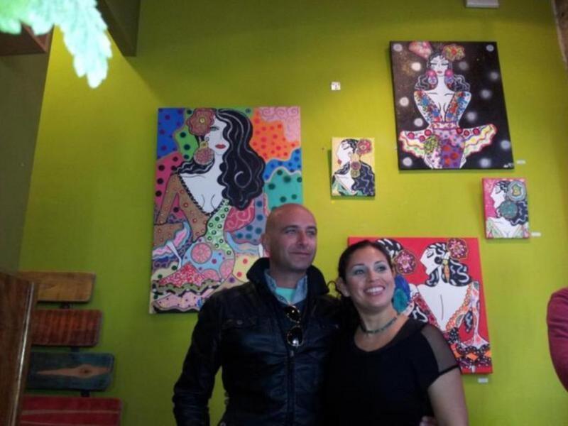 Dos personas posan frente a una pared amarilla con cuadros de arte pop coloridos.