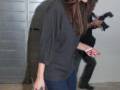A una mujer con cabello largo y lacio, llevando un suéter gris oscuro, pantalones vaqueros azules y una bolsa de mano verde. Está sosteniendo un teléfono móvil con la otra mano. La escena parece ser al aire libre, posiblemente en una zona urbana con paredes de hormigón.