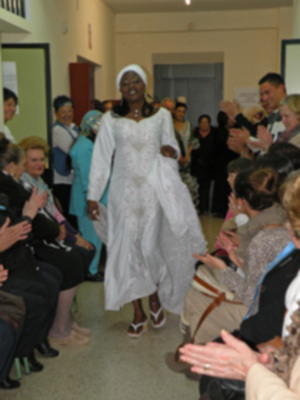 Una mujer vestida con un atuendo blanco y largo camina entre una audiencia de personas que aplauden. La imagen parece ser de un evento formal o ceremonial, posiblemente una boda o ceremonia religiosa.