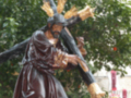 Una figura de madera policromada representa a Jesús con una cruz sobre la cabeza, rodeado por velas y flores. La escena se desarrolla en un entorno natural con árboles al fondo, sugiriendo una celebración religiosa o procesión.