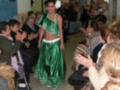 Una mujer en traje verde brillante y falda larga, con un vestido de gala, camina entre una audiencia entusiasta que aplaude y toca. La escena parece ser un evento formal o una competencia de baile, con una decoración interior simple y una puerta visible en el fondo.