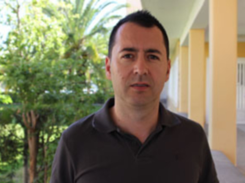 Un hombre con cabello corto y una camisa de polo negra, posando frente a un edificio amarillo con ventanas blancas. El fondo muestra árboles verdes y una sombra de un palmera en el cielo.