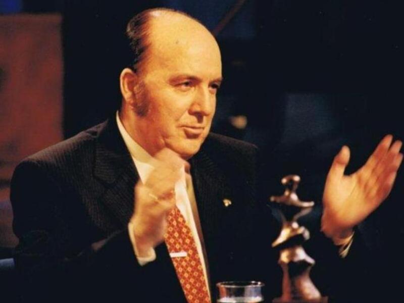 Un hombre en traje hablando con gesto de discusión, frente a un fondo oscuro.