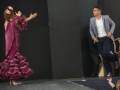 Una bailarina de flamenco en acción, con un vestido de flamenca de color rosa y blanco, mientras un bailarín observa en el fondo.