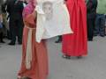 Una persona vestida con una túnica roja y un manto beige sostiene un lienzo blanco con una imagen de Jesús crucificado. La escena parece ser parte de una procesión religiosa en un ambiente urbano con otros participantes y observadores.