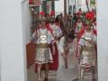 Un grupo de personas vestidas con trajes tradicionales, posiblemente de una celebración o fiesta, caminan por un arco blanco. La imagen muestra detalles como el sol y algunas estructuras urbanas en el fondo, sugiriendo un entorno urbano histórico.