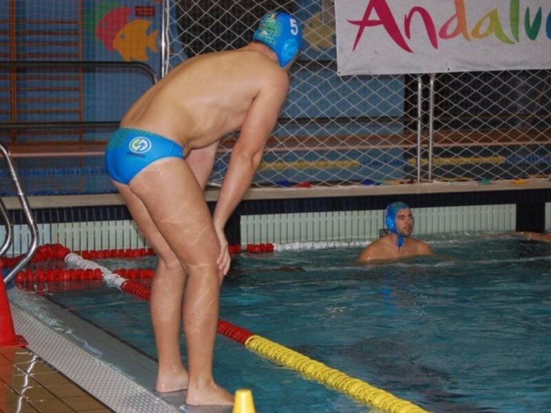 Un nadador con una camiseta azul y un casco de natación se prepara para nadar en una piscina. En el fondo, otro nadador está listo para nadar y un cartel con la palabra "Andalucía" se puede ver en el borde de la piscina.
